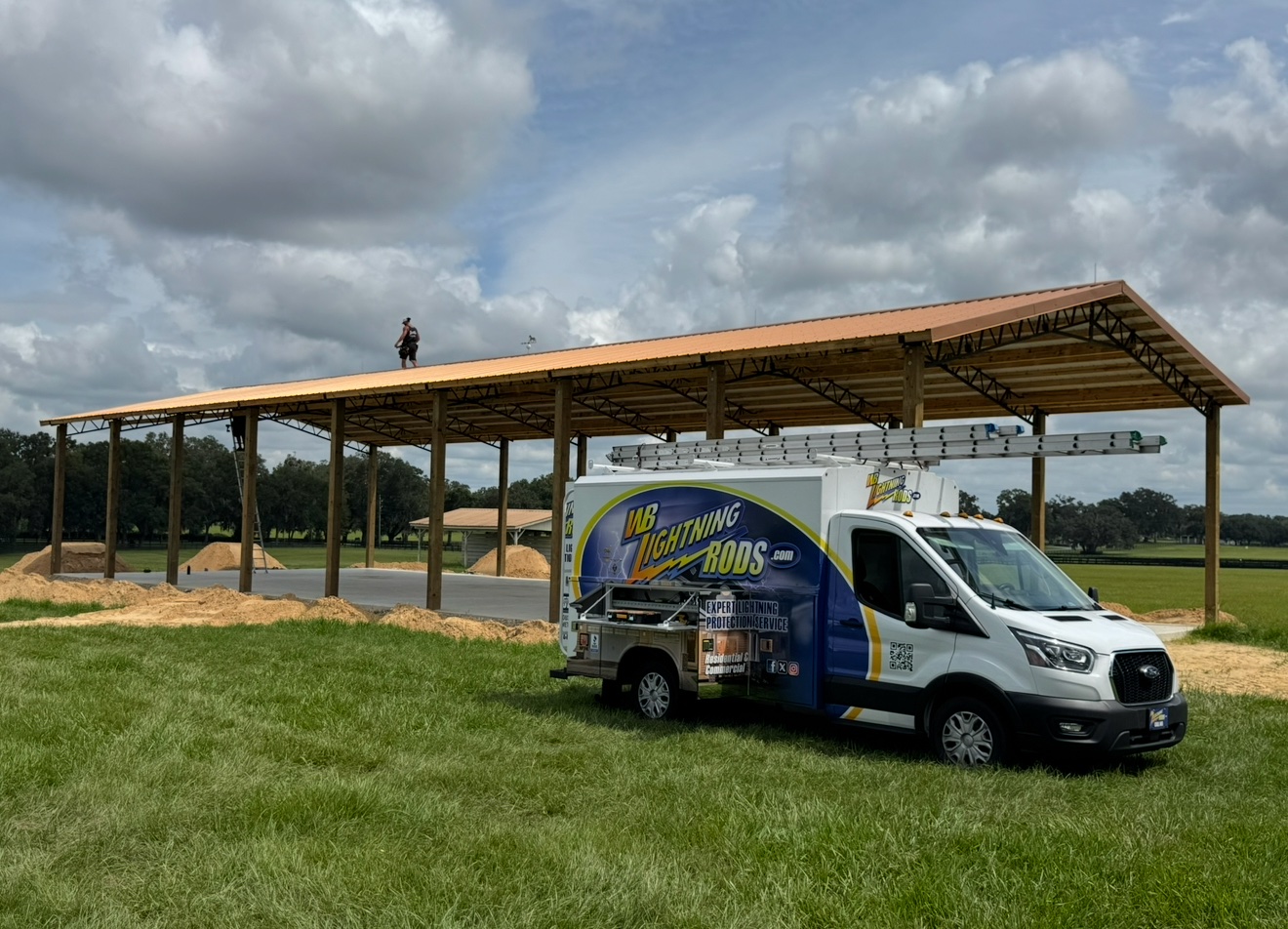 WB Lightning Rods van parked in front of a large open pole barn with a copper-colored metal roof, with a crew member on the roof